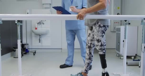 白人男性理学療法士がノートを作成する一方で 義肢のある白人女性患者は 病院での理学療法セッション中に平行棒の間を歩く スローモーション — ストック動画