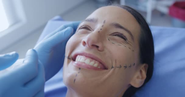 一位白种人女性病人躺在诊所的手术台上笑着 她的脸上挂着整容的图样 一位整容医生的戴着手套的手指向她的脸 动作缓慢 — 图库视频影像