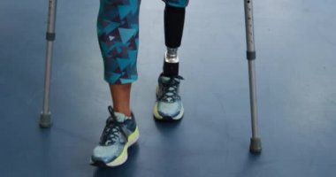 Spor salonundaki koltuk değnekleriyle yürüyen protez bacaklı, beyaz bir kadının ön görüntüsü. 