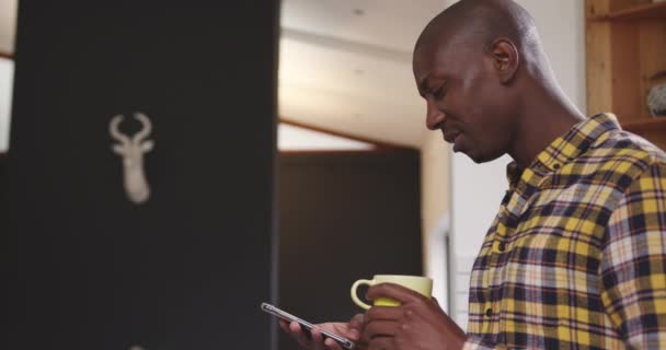 一个非洲裔美国人穿着格子衬衫站在厨房里 拿着一杯咖啡 拿着智能手机 面带微笑 慢吞吞地走起路来 — 图库视频影像