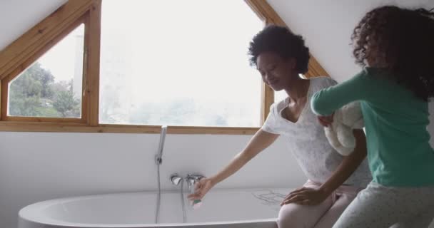 一个非洲裔美国妇女和她年幼的女儿在浴室里 母亲坐在浴缸边洗澡 女儿站在她旁边抱着泰迪熊 动作缓慢 — 图库视频影像