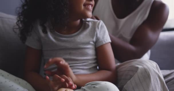 在客厅里 一个非裔美国男人和他的小女儿坐在沙发上 一个女孩正在看电视 一个女孩在笑 而她的父亲在梳理她的头发 动作缓慢 — 图库视频影像
