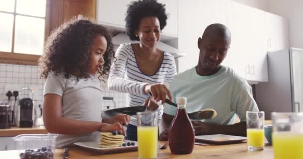 アフリカ系アメリカ人の少女と彼女の父親の朝の台所の家の正面図は キッチンの島に座って 母親はフライパンからパンケーキを提供して後ろに立って — ストック動画