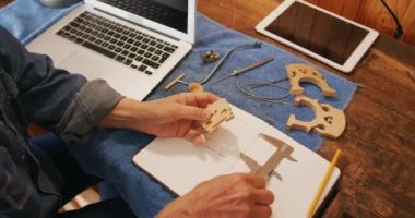 Atölyesindeki atölyesinde çalışan üst düzey bir beyaz kadın Luthier 'in yüksek açılı görüntüsü, keman köprüsünü ölçüyor ve önündeki atölyesinde dizüstü bilgisayar, tablet bilgisayar ve aletlerle not alıyor.
