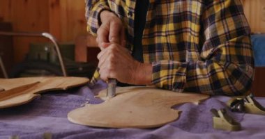 Üst düzey bir beyaz kadın Luthier 'in, atölyesindeki bir tezgahta keman üzerinde çalışırken, arka plandaki duvarda asılı aletlerle arka panelin ahşabını şekillendirmek için keski kullanırken ön görüntüsü..