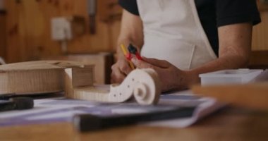 Beyaz önlük giyen, atölyesindeki bir tezgahta keman yapan, elinde pusula tutan ve kalemle çizim yapan beyaz önlüklü beyaz bir kadın Luthier 'in ön tarafı.
