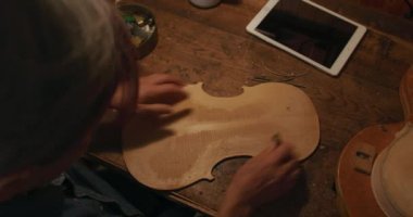  Kafkas kadın Luthier atölyesinde bir kemanın arkasında çalışıyor, onu tutuyor ve eserini inceliyor, önünde bir tablet bilgisayar ve arka planda duvarda asılı aletlerle..