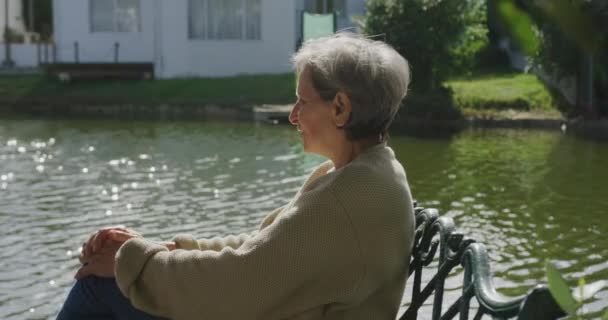 一位白种人的老年妇女坐在河边的长椅上 在阳光下悠闲自在地欣赏风景 动作缓慢 — 图库视频影像