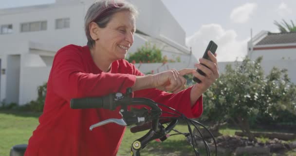 一位白种人的老年妇女穿着一件红色毛衣 坐在街上的自行车上 在阳光下微笑 动作缓慢 侧视近景 — 图库视频影像