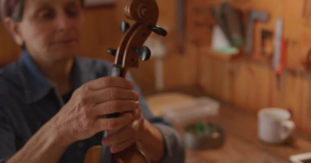 フロントビュー閉じるのシニア白人女性Luthier巻き弦上のチューニングペグ上の彼女のワークショップで完成したバイオリン 首を検査し スクロール — ストック動画