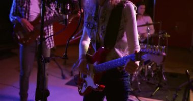 Sarışın, kıvırcık saçlı beyaz kadın gitarist, karışık ırktan erkek gitarist ve beyaz bir erkek baterist, hepsi de kulaklık takıyor ve bir kayıt stüdyosunda birlikte şarkı söylüyorlar. Müzisyenler bir şarkı üzerinde çalışıyor.