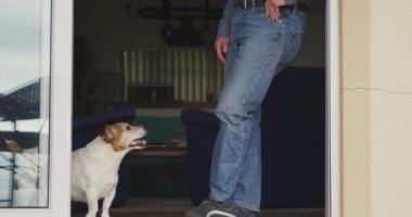 Yaşlı, beyaz bir adamın evde dinlenirken, ayakta dikilirken, kapı eşiğine yaslanırken, evcil köpeğine bakarken, ağır çekimde...