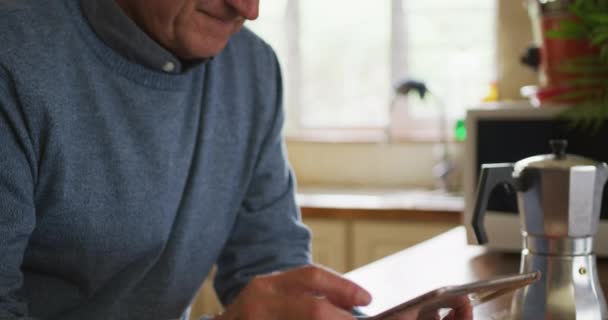 一位年长的白人男子在家里放松下来 坐在厨房的柜台旁 用平板电脑 慢吞吞地微笑着 侧视近景 — 图库视频影像