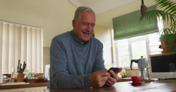 一个年长的白人男子在家里放松下来 坐在厨房的柜台前 用智能手机 慢吞吞地笑着 — 图库视频影像