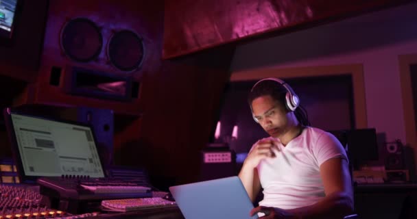 混合レースの男性サウンドエンジニアのフロントビューは ヘッドフォンを着用し ラップトップコンピュータを使用して録音スタジオのミキシングデスクで座って作業しています サウンドエンジニアとして楽曲制作に取り組む — ストック動画