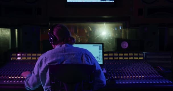 録音スタジオのミキシングデスクに座っているポニーテールを持つ白人男性サウンドエンジニア 彼の頭の後ろに手を置き ヘッドフォンで聞いて コンピュータ画面を見て ライブ録音室で演奏するバンド — ストック動画