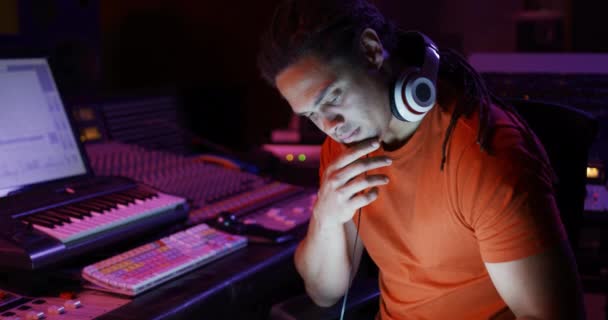 一个混血的男声工程师坐在录音室里的一个混音桌旁 脖子上戴着耳机 侧视近景 负责创作歌曲的音响工程师 — 图库视频影像