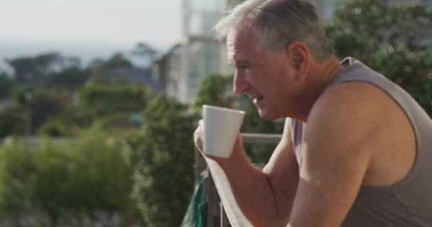 旁边是一位身穿背心 站在阳台上晒太阳 早上喝一杯咖啡 慢动作的白人老人在家里放松的侧影 — 图库视频影像