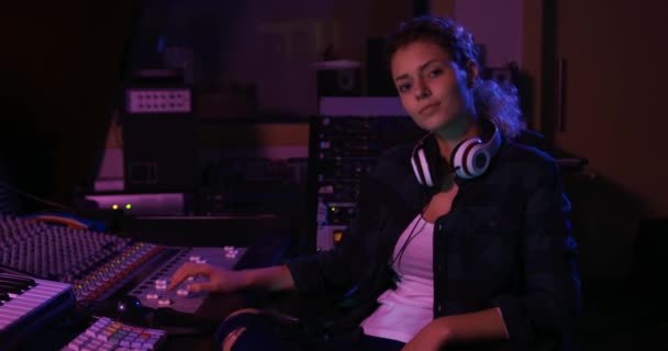 一个年轻的白人女声工程师坐在录音室里的一张混合桌子前 脖子上戴着耳机 对着摄像机笑着的画像 负责创作歌曲的音响工程师 — 图库视频影像