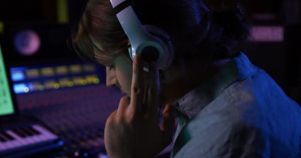 サイドビューは ヘッドフォンを装着した白人男性サウンドエンジニアの近くにあり コンピュータインターフェースを使用してレコーディングスタジオのミキシングデスクで作業しています サウンドエンジニアとして楽曲制作に取り組む — ストック動画