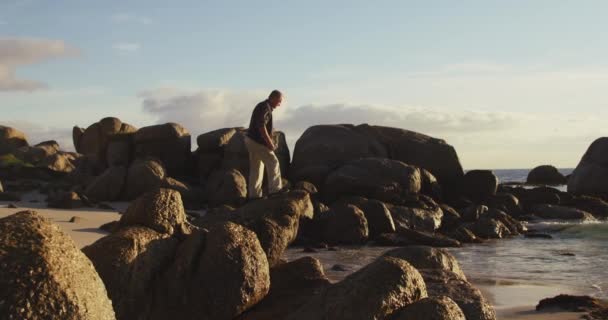 ビーチで岩の上を一人で歩いている上級白人男性の側のビューでは 海を見て スローモーション — ストック動画