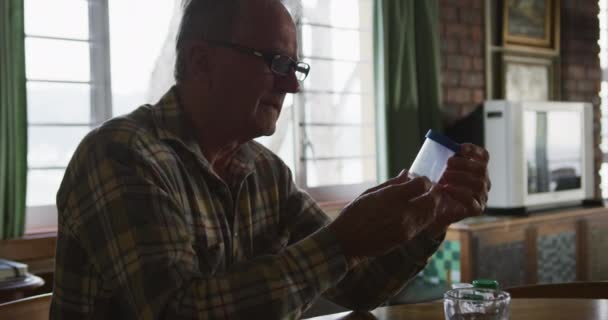 一个白人老人呆在家里 戴着眼镜坐在桌旁 拿着一瓶药片 阅读标签 慢动作的侧视图 — 图库视频影像