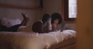 Side view of a young mixed race woman with afro hair and her African American male partner, lying on their bed leaning on their elbows smiling and watching a laptop computer together at home