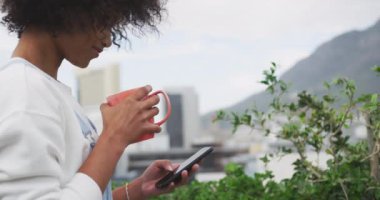 Side view close up of a young mixed race woman with afro hair standing outside on a roof terrace in the city drinking a cup of coffee and using smartphone smiling, slow motion
