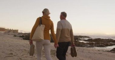 Rear view of a happy senior Caucasian couple enjoying time in nature together, holding hands, walking barefoot alongside beach at sunset, slow motion