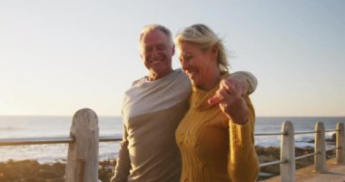 Front view of a happy senior Caucasian couple enjoying time in nature together, smiling and embracing, walking alongside beach at sunset, slow motion
