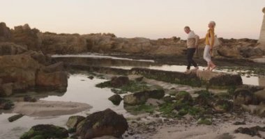 Side view of a happy senior Caucasian couple enjoying time in nature together, holding hands, walking barefoot alongside beach on rocks at sunset, slow motion