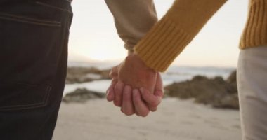 Rear view mid section of a senior Caucasian couple enjoying time in nature together, walking on a beach at sunset, holding hands, slow motion