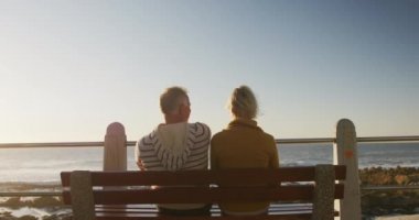 Rear view of a senior Caucasian couple enjoying time in nature together, sitting on a bench by the sea, embracing and admiring the view, slow motion