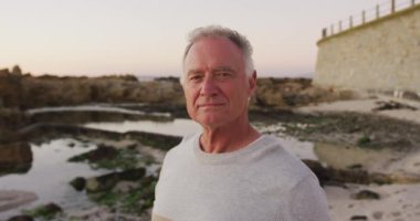 Portrait of a happy senior Caucasian man enjoying time in nature, smiling to camera, standing by the sea on a beach at sunset, slow motion