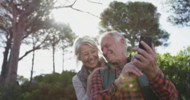 Front view of a senior Caucasian couple enjoying time in nature together, standing in forest during a hike, smiling and taking a selfie with smartphone, slow motion