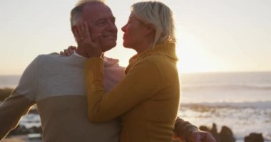 Front view of a happy senior Caucasian couple enjoying time in nature together, smiling and embracing, kissing, standing by a balustrade alongside beach at sunset, slow motion