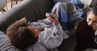 Karışık ırktan olan bir çiftin yüksek açılı yan görüntüsü. Evde birlikte vakit geçiriyorlar. Kanepede uzanıyorlar ve oturma odasında, akıllı telefon ve dizüstü bilgisayar kullanıyorlar.