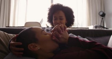 Yan görüntüde, melez bir kadın çiftin evde vakit geçirmesinin keyfini çıkarırken, oturma odasında bir koltukta el ele tutuşurken, bir kadın diğer kadının kucağında dinlenirken, ağır çekimde.