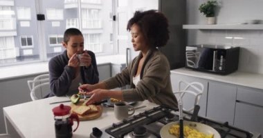 Yüksek açılı, melez bir kadın çiftin evde birlikte vakit geçirmesini mutfak masasında kahvaltı etmesini pencereden görüldüğü bir binayı ağır çekimde izlemesini.