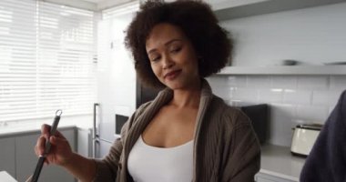 Melez bir kadın çiftin evde birlikte vakit geçirmesinin yan görüntüsü. Mutfakta kahvaltı hazırlıyorlar. Karıştırıyorlar, birbirlerine bakıyorlar ve gülümsüyorlar.