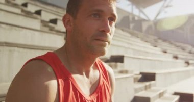 Boş bir spor stadyumundaki beyaz erkek sporcunun yan görüntüsü. Yarışmadan önce oturup konsantre oluyor. Yavaş çekim.