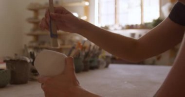 Beyaz tenli, kısa saçlı genç bir çömlekçinin çalışma masasında oturup çömlek atölyesinde jöleyle fırçalarkenki görüntüsü, yavaş çekim.