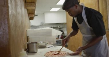 Side view of a mixed race male cook working in a busy pizza restaurant kitchen, placing tomato sauce over pizza dough before cooking, female cook walking in the background