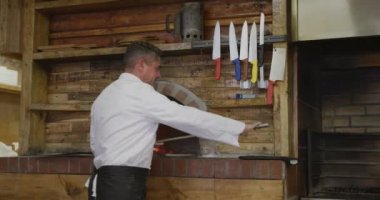 Portrait of a happy Caucasian male chef working in a busy pizza restaurant kitchen, putting a pizza peel on a shelf, smiling to camera and crossing his arms, pizza oven in the background
