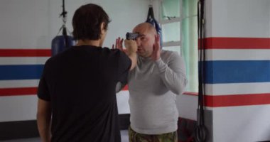 Krav Maga Training. Front view close up of a bald Caucasian male instructor giving self defence training in a boxing gym demonstrating a gun defense to a Caucasian man, while another man looks on, slow motion