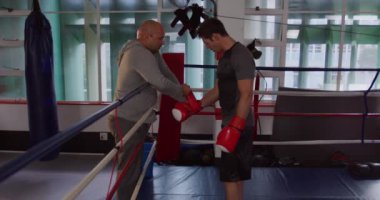 Side view of a Caucasian male boxer standing in a boxing ring while a bald Caucasian male trainer is wrapping his glove, slow motion