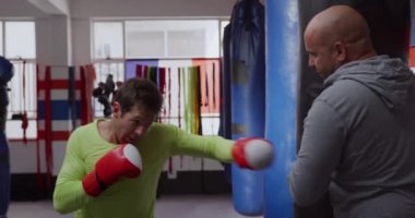 Side view of a Caucasian male boxer punching a punchbag hold by a bald Caucasian male trainer in a boxing gym, slow motion
