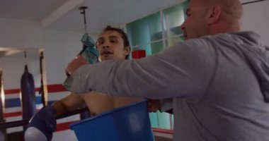 Front view close up of a mixed race male boxer drinking water gave by his bald Caucasian male instructor in a boxing ring, slow motion