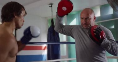 Side view of a mixed race male boxer practicing a punch against a pad held by a bald Caucasian male trainer in a boxing gym, slow motion