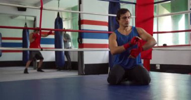 Front view of a mixed race male boxer wrapping his hands with boxing bands kneeling in a boxing ring at a boxing gym, with a trainer and another male boxer training in the background, slow motion